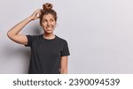 Small photo of Horizontal shot of confused unaware European woman with hair bun scratches head clenches teeth and looks aside tries to recall something in mind wears black t shirt isolated over white background