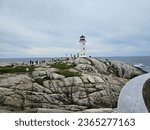 Small photo of Peggy's Cove, NS, CAN, August 15, 2023 - The Peggy's Cove lighthouse with some tourists standing on the rocks around it on a cloudy summer day.