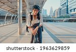 Small photo of Asian businesswoman go to work at office stand and smiling wear backpack look at camera with bicycle on street around building on a city. Bike commuting, Commute on bike, Business commuter concept.