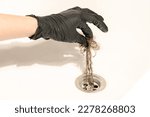 Small photo of A bathroom drain clogged with dirty hair and slime. A female hand in a black latex glove pulls a long messy tuft of hair out of the drain