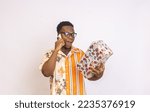 Small photo of A Young happy eloquent African man wearing transparent glasses, holding a gift box, and making a phone call in front of a white background.