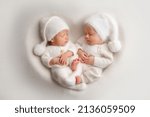 Small photo of Tiny newborn twin boys in white bodysuits on a white background in white caps. Newborn twins sleep next to their brother on the background of the heart. Two newborn twin boys hugging each other.