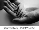 Small photo of A parent holds the hand of a newborn child. The small hand of the son with tiny fingers lies on the palm of the father. The concept of a happy family. Family hereditary ties. Black and white photo.