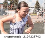 Small photo of A little joyful girl bathes in a fountain in the summer heat, squeals merrily and runs away from high jets of water, a lot of splashes.