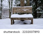A snow-covered wooden bench in a winter park. Next to the bench is a round trash can.