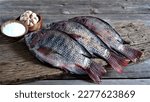 Small photo of Tilapia. Fresh Tilapia fish on board with a cup of salt, and garlic. Oreochromis Niloticus. Freshwater Fish. In Indonesia also known as Ikan Nila or Mujair. Farmed Fish. Tribe Tilapiine cichlid.