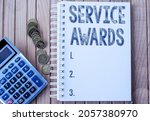 Small photo of Text showing inspiration Service Awards. Business approach Recognizing an employee for his or her longevity or tenure Plain Spiral Notebook With Pen Beside Calculator And Coins On Table.