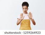 Small photo of Calm down, put gun down. Intense worried and nervous scared african-american woman with curly hair in yellow overalls, raising palms, asking to relaxed and chill, trying comfort depressed friend
