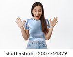 Image of surprised brunette girl smiles, looks in awe down, raise hands and rejoice, found sale promo below, staring at bottomg advertisement, standing against white background