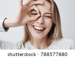 Close up portrait of beautiful joyful blonde Caucasian female smiling, demonstrating white teeth, looking at the camera through fingers in okay gesture. Face expressions, emotions, and body language