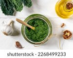 Small photo of Homemade kale pesto sauce served in a glass bowl over white wooden surface with basic ingredients - kale leaves, walnuts, parmesan, garlic and olive oil