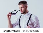 Stylish and fashionable man in classical wear wearing black sunglasses while standing against gray background. Elegant man in white shirt and suspenders posing in studio. Men's classic eyewear fashion