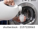 Against background of drum of steel-colored washing machine, woman pours liquid washing gel into plastic cap. A girl in a white T-shirt carefully pours a transparent conditioner for flattening laundry