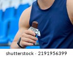 Small photo of Muscular athlete snacks on a protein bar while resting after a workout at the stadium. Man in a blue sports jersey rests in the stand for fans, eating a chocolate bar.