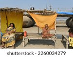 Small photo of Prayagraj,UP,01 14 2023:A man with bare body meditating with folded hands in winters at river Ganges bank. On next wooden cot, a lady sitting and waiting, behind is platoon bridge.