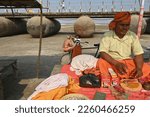 Small photo of Prayagraj,UP,01 14 2023: Hindu priest waits devotees to put them a yellow mark on forehead. Behind is platoon bridge over river Ganges.