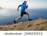 Small photo of male runner running uphill trail on edge in blue jacket and black tights, background of sky and sea