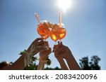 Women hands toasting with aperol spritz cocktails on summer party on blue sky background, copy space. Event celebration concept. Traditioanal italian aperitif.