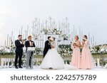 Front view of wedding ceremony outdoors, man in suits kissing woman in white puffy dress, while standing with bridesmaids and groomsmen near floral arch.