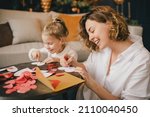 Small photo of Mother and little daughter making Valentine's day cards using color paper, scissors and pencil, sitting by the table in cozy room