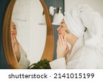 Beautiful young woman with perfect skin wearing white bathrobe and towel on head after shower, making face massage using a jade face roller with natural quartz stone in bathroom.