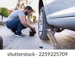 Small photo of young man loosening a nut of his car tire, changing a flat tire on the road, car jack being used. young man loosening a nut of his car tire, changing a flat tire on the road