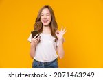 Cheerful Asian woman holding smartphone and shows ok sign on light yellow background.