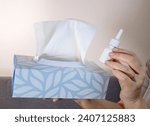 Small photo of Close up of woman's hands holding a tissue box and a nose spry bottle, white blurry background