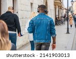 The back of a black man in a denim jacket, jeans with black wireless headphones among passers-by on the street on a sunny day