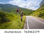 Small photo of Active man with a red rucksack is walking afoot along a road in the mountains of Scotland, summer time.