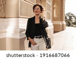 Happy girl with short hair in light trauswrs and dark jacket with handbag smiling outdoors. Modern lady in glasses posing in city..