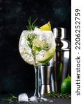 Small photo of Gin tonic lime alcoholic cocktail drink with dry gin, rosemary, tonic and ice in big wine glass. Black bar counter background, steel bar tools