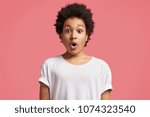 Small photo of Curly little African boy looks with bated breath and shocked expression, keeps mouth widely opened, being afraid of something, wears casual white t shirt isolated over pink background. Concerned child