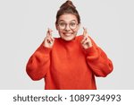 Lucky beautiful Caucasian female crosses fingers , hopes all wishes come true, wears oversized red sweater and spectacles, isolated over white background. People, body language and happiness