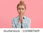 Shot of miserable young female keeps palms together near mouth, has pleading expression, light hair combed in bun, wears denim fashionable jacket, poses against pink background. Please, help me!