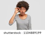 Small photo of Portrait of astonished lovely African America female with crisp hair, looks with bated breath, being terrified by what she sess, keeps hand on spectacles, isolated on white studio background