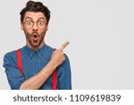 Surprised male hipster, keeps mouth opened, being stupefied as advertises something, wears fashionable shirt and red suspenders, indicates with fore finger at upper right corner. Omg concept