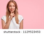 Indoor shot of beautiful female looks mysteriously aside, has intriguing look, asks to be quiet, dressed in casual clothes, stands against pink background with blank copy space for your advertisement
