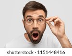 Close up portrait of stunned bearded young guy drops jaw, has bugged dark eyes, sees something unbelievable and surprising, has eyewear, isolated on white background. People, emotions concept