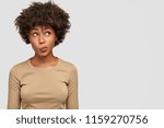 Small photo of Young lovely female has dark skin, clueless and unaware expression, purses lips as being questionned, looks with puzzlement upwards, isolated over white background with copy space on right side