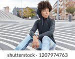 Carefree ethnic young woman wears casual sweatshirt with hood, leggings sits on road against urban background, focused into distance, thinks about favourite sport. People and recreation concept