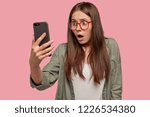 Small photo of Shocked woman sees embarrasing picture in screen of smart phone, stares with amazement at camera, wears spectacles, has bated breath, isolated over pink background. Girl talks via video chat