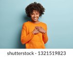 Touched beautiful smiling Afro American girl keeps palms on heart, expresses appreciation, pleasure and gratitude, wears orange sweater, models over blue background. Confession in love concept.