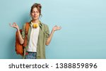 Photo of relaxed student keeps hands in mudra gesture, keeps eyes closed, listens peaceful music, headphones on neck, wears shirt and jeans, has rucksack, isolated on blue background with free space