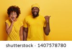 Small photo of Studio shot of surprised woman with bated breath, stands closely to dark skinned guy who points aside on blank space, see something awesome, wear casual yellow t shirts, express shock and joy
