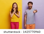 Small photo of Photo of shocked couple gaze straightly at camera with bated breath, happy unshaven man in casual wear points at something, stands over purple background, impressed woman looks with excitement