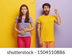 Hungry dark haired woman touches stomach, wants to eat something tasty, wears purple t shirt and pink trousers, impressed unshaven man in casual wear points index finger above, shows direction up