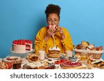 Small photo of Gluttony and overeating concept. Upset crying ethnic woman eats piece of cake reluctantly, sits at table with many desserts, isolated over blue wall, feels hungry and greedy, wears yellow jacket