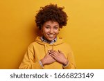 Portrait of dark skinned friendly woman makes gratitude gesture, expresses thankfullness for received compliment, wears hoodie, isolated over yellow background, got surprise or praise, being grateful