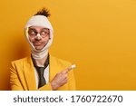 Small photo of Funny positive man injured and has bruises on face, beaten by cruel people, wears black tie and yellow jacket, indicates away on empty space, fell from staircase at home. Accident and pain concept